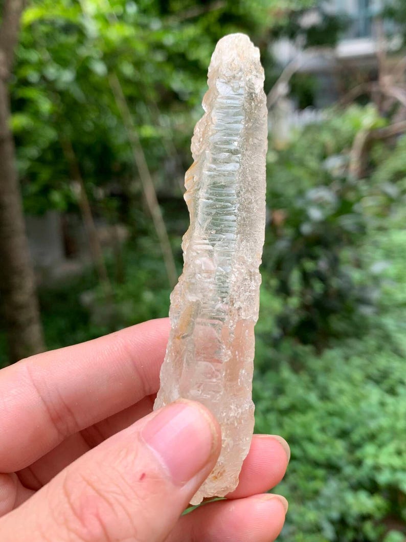 Puede incluir: Una formaci&oacute;n cristalina transl&uacute;cida y clara, con una superficie rugosa y texturizada. El cristal se sostiene en una mano sobre un fondo verde borroso. Es un esp&eacute;cimen mineral natural.