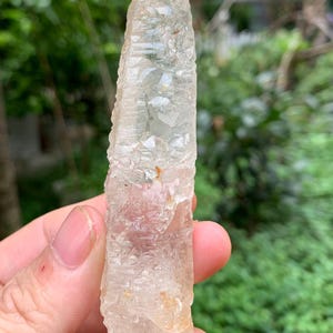Puede incluir: Un cristal claro y alargado con una superficie rugosa y texturizada. El cristal tiene una apariencia p&aacute;lida y transl&uacute;cida con sutiles variaciones de color. El cristal se sostiene contra un fondo verde borroso.