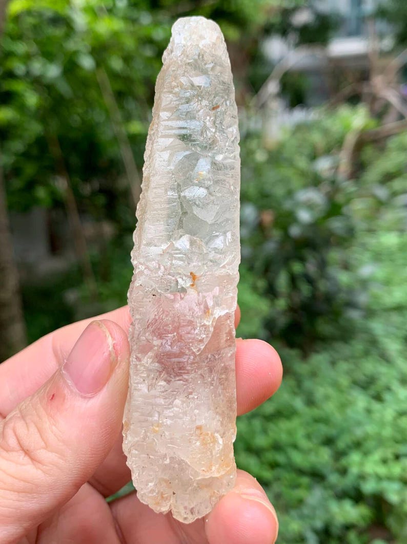 Puede incluir: Un cristal claro y alargado con una superficie rugosa y texturizada. El cristal tiene una apariencia p&aacute;lida y transl&uacute;cida con sutiles variaciones de color. El cristal se sostiene contra un fondo verde borroso.