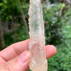 Puede incluir: Un cristal claro y alargado con una superficie rugosa y texturizada. El cristal se sostiene en una mano, con un fondo verde borroso. El cristal tiene un color amarillo p&aacute;lido y claro.