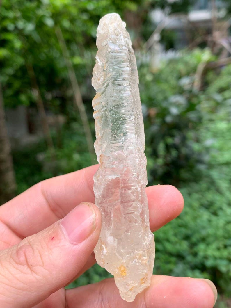 Puede incluir: Un cristal claro y alargado con una superficie rugosa y texturizada. El cristal se sostiene en una mano, con un fondo verde borroso. El cristal tiene un color amarillo p&aacute;lido y claro.