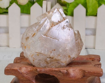 May include: A large, clear quartz crystal with a unique, faceted shape. The crystal is resting on a wooden stand.