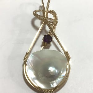 May include: A handcrafted gold pendant featuring a large, iridescent, white, coin-shaped pearl. The pendant is accented with a small, round, purple gemstone and intricate wire wrapping.