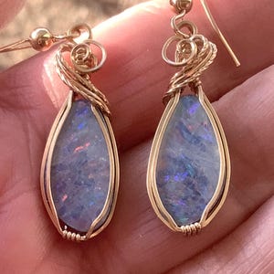 May include: A pair of gold-toned wire-wrapped earrings. Each earring features a teardrop-shaped, iridescent opal stone. The opals display shades of blue, purple, and red. The earrings have a delicate, handcrafted appearance, with intricate wire detailing.