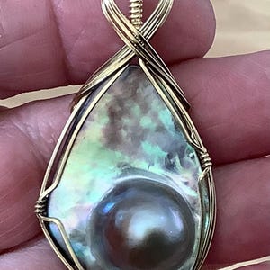 May include: A pear-shaped pendant featuring a large, iridescent pearl set within a gold wire frame. The pearl has a dark, lustrous surface with hints of blue and green. The gold wire is intricately wrapped, forming a decorative bail.