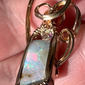 May include: A wire-wrapped pendant featuring a rectangular opal with iridescent flashes of pink, blue, and green. The stone is encased in polished gold-colored wire, which forms intricate swirling patterns and a small loop at the top for hanging.