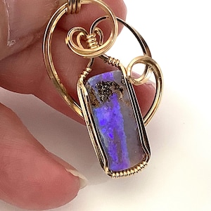 May include: A pendant featuring a rectangular boulder opal with vibrant purple and blue flashes, encased in a gold-colored wire-wrapped setting. The wire forms decorative swirls and loops around the stone, creating a frame for the gemstone.