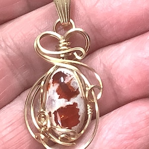 May include: Gold-toned pendant with an oval-shaped, multi-colored stone. The stone features a mix of white and reddish-brown hues. The pendant is intricately designed with wire wrapping and decorative swirls, creating a unique jewelry piece.