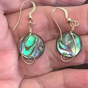 May include: A pair of gold-colored earrings with iridescent, round abalone shell pendants. The earrings have a hook design and feature a wire-wrapped spiral detail. The abalone shells display shades of green, blue, and purple.