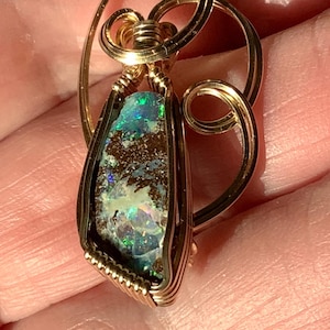 May include: A pendant featuring a raw, dark-toned boulder opal with vibrant green and blue flashes, encased in a decorative gold-colored wire-wrapped setting with swirling loops at the top, creating a detailed and intricate metal frame.