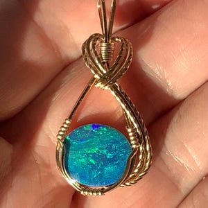 May include: A round, iridescent blue and green opal gemstone set in a gold wire-wrapped pendant. The gold wire forms a teardrop-shaped frame around the stone, with a decorative coiled bail at the top for attaching to a necklace chain.