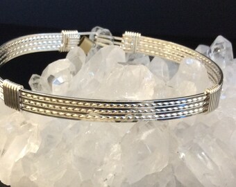 May include: A silver-colored bangle bracelet with a decorative design. The bracelet features multiple parallel bands with a textured pattern. The clasp is visible at the top. The bracelet is displayed on a bed of clear quartz crystals.