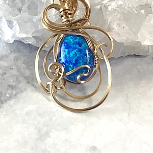 May include: A gold wire wrapped pendant featuring a blue opal gemstone. The pendant has a delicate, intricate design with swirling wire.