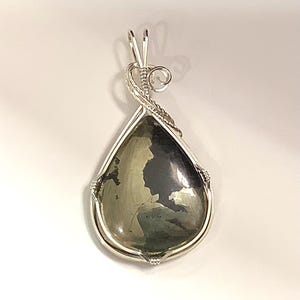 May include: A teardrop-shaped pendant with a silver wire wrap. The pendant features a polished, iridescent stone with shades of gray, black, and gold. The silver wire forms a decorative bail at the top.