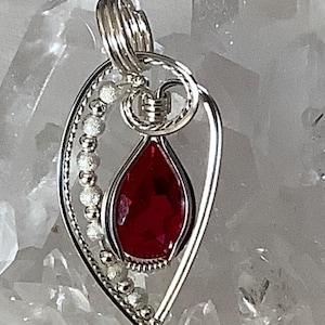 May include: A silver wire wrapped pendant featuring a teardrop-shaped red gemstone. The pendant is adorned with small silver beads and has a delicate wire design.