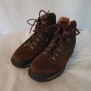 Puede incluir: Un par de botas de senderismo de ante marrón y tela verde oscuro con diseño de caña alta, suelas de goma negra, ojales metálicos en forma de D y cordones marrones. Las botas cuentan con un cuello acolchado y una puntera reforzada.