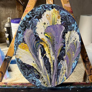 May include: Oval abstract painting featuring stylized flowers in white, purple, and yellow against a dark blue background. The artwork is mounted on a wooden easel, showcasing a fluid paint technique. The painting's dimensions are not visible.