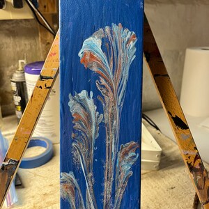 May include: An abstract painting on a rectangular canvas. The background is a deep blue, with a floral design in shades of white, light blue, and rust. The artwork is displayed on a wooden easel, showcasing the textured brushstrokes.