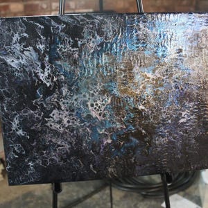 May include: Abstract acrylic painting on canvas, displayed on a black easel. The artwork features a textured surface with a blend of dark blues, blacks, white, and gold. The painting has a fluid, marbled appearance.