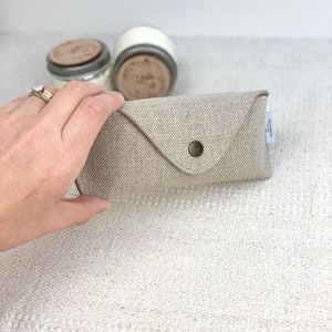 May include: A beige linen eyeglass case with a button closure. The case is made of a soft, natural fabric and has a simple, elegant design.