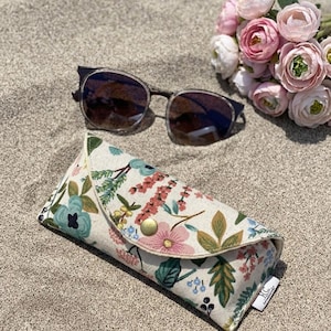 Könnte beinhalten: Sonnenbrille mit dunklen Gläsern und einem dünnen, dunklen Rahmen liegen auf Sand neben einem Brillenetui mit Blumenmuster und einem Strauß rosa Blumen. Das Brillenetui hat ein Muster aus rosa, blauen und grünen Blumen und einen goldenen Druckknopfverschluss.