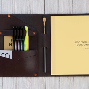 May include: A brown leather notebook cover with a built-in pen holder and a pocket for cards. The cover is open to reveal a yellow notebook with the text "HOBONICHI TECHO 2020 cousin" printed on the front.