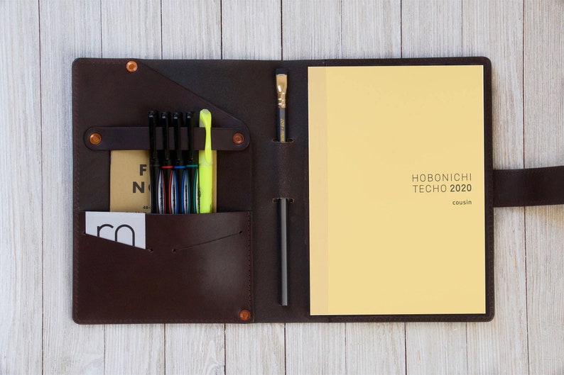 May include: A brown leather notebook cover with a built-in pen holder and a pocket for cards. The cover is open to reveal a yellow notebook with the text "HOBONICHI TECHO 2020 cousin" printed on the front.
