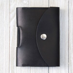 K&ouml;nnte beinhalten: Eine schwarze Ledergeldb&ouml;rse mit einem gebogenen Klappverschluss, der mit einem silbernen Druckknopf gesichert ist. Die Geldb&ouml;rse ist rechteckig und scheint geschlossen zu sein. Der Hintergrund ist eine hellfarbige Holzoberfl&auml;che.
