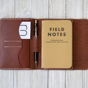 K&ouml;nnte beinhalten: Eine braune Leder-Notizbuchh&uuml;lle mit einem Field Notes Notizbuch, einem Stift und einer Karte. Die H&uuml;lle ist offen und zeigt die Innentaschen und das Notizbuch. Die H&uuml;lle wird mit einem Druckknopf verschlossen.