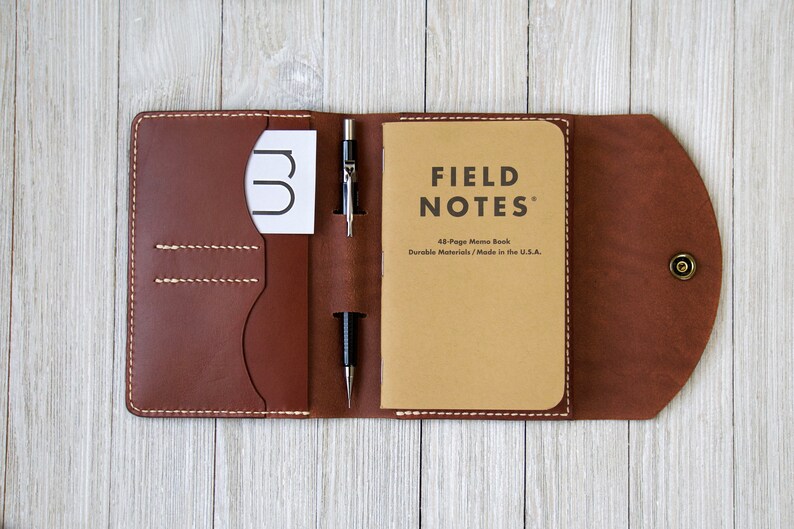 K&ouml;nnte beinhalten: Eine braune Leder-Notizbuchh&uuml;lle mit einem Field Notes Notizbuch, einem Stift und einer Karte. Die H&uuml;lle ist offen und zeigt die Innentaschen und das Notizbuch. Die H&uuml;lle wird mit einem Druckknopf verschlossen.