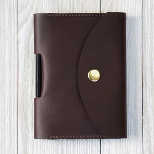 K&ouml;nnte beinhalten: Eine dunkelbraune Leder-Notizbuchh&uuml;lle mit einem goldenen Knopfverschluss. Die H&uuml;lle hat eine Stifthalterung an der Seite. Das Notizbuch ist geschlossen und liegt auf einer hellen Holzoberfl&auml;che.