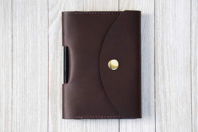 K&ouml;nnte beinhalten: Eine dunkelbraune Leder-Notizbuchh&uuml;lle mit einem goldenen Knopfverschluss. Die H&uuml;lle hat eine Stifthalterung an der Seite. Das Notizbuch ist geschlossen und liegt auf einer hellen Holzoberfl&auml;che.