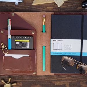 May include: A brown leather notebook cover with a built-in pocket for a pen and a small notebook. The cover has a black Moleskine notebook inside with the text "Classic Collection Dotted Notebook Carnet à grille de pointillés" on the front.