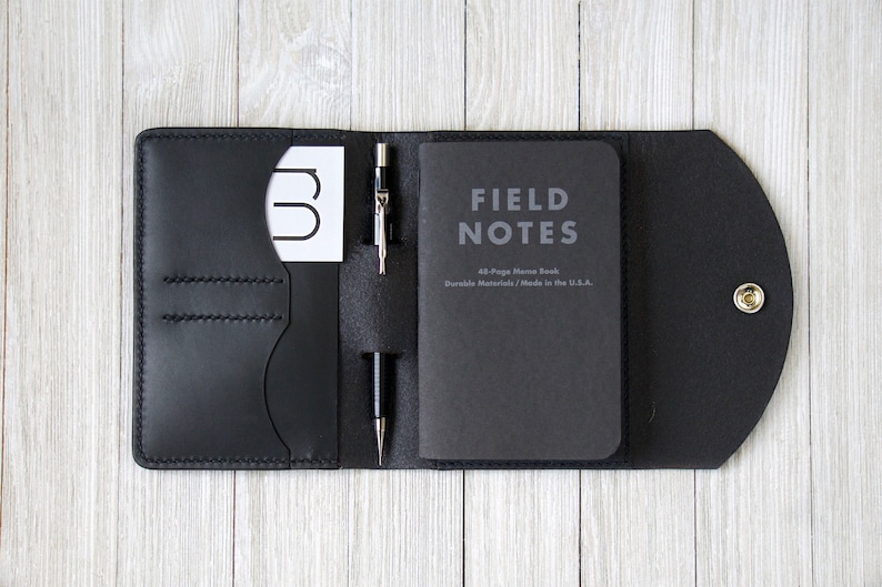 K&ouml;nnte beinhalten: Schwarze Leder-Notizbuchh&uuml;lle mit Druckknopfverschluss, die ein Field Notes-Notizbuch, eine Visitenkarte und zwei Stifte enth&auml;lt. Die H&uuml;lle ist ge&ouml;ffnet und zeigt den Inhalt. Auf dem Notizbuch steht "FIELD NOTES".