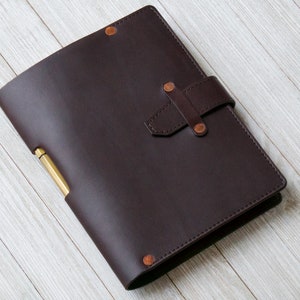 May include: A dark brown leather bound journal with a brass pen holder and a leather strap closure. The journal is on a white wooden surface.