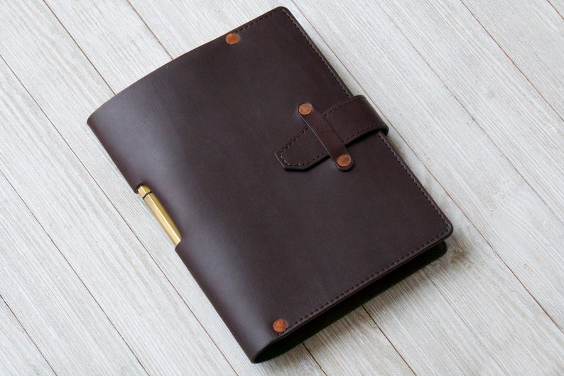 May include: A dark brown leather bound journal with a brass pen holder and a leather strap closure. The journal is on a white wooden surface.