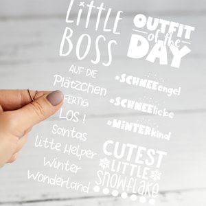 Könnte beinhalten: Ein klares Schild mit weißem Text, gehalten von einer Hand. Auf dem Schild steht "Little Boss Outfit of the Day" mit weiteren Sätzen wie "Cutest Little Snowflake" und "Winter Wonderland". Der Hintergrund ist eine verschwommene, hellfarbige Holzoberfläche.