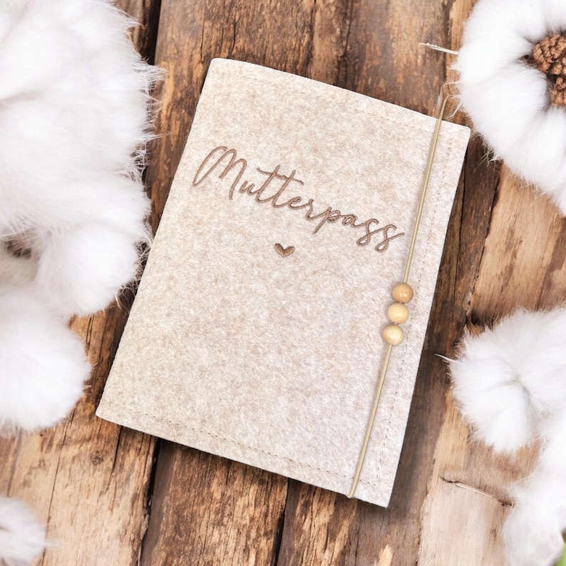 May include: A beige felt cover with the word "Mutterpass" in a script font and a small heart. A string with three wooden beads is attached to the side. The cover is on a wooden surface.