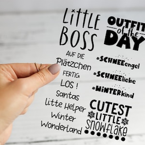 Könnte beinhalten: Ein klarer Aufkleber mit schwarzem Text, gehalten von einer Hand. Der Text enthält Sätze wie "Little Boss", "Outfit of the Day" und "Cutest Little Snowflake". Weitere Wörter sind "Winter" und "Wonderland".
