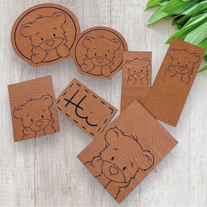 Könnte beinhalten: Eine Sammlung brauner Lederaufnäher mit einem Cartoon-Bär-Design. Die Aufnäher haben verschiedene Formen, darunter Kreise, Rechtecke und ein Quadrat. Einige Aufnäher haben den Text "Hi" aufgenäht. Der Bär ist in Schwarz gezeichnet.