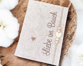 Op de afbeelding: Een beige vilten kaart met de woorden "Liebe im Bauch" in bruin geborduurd. De kaart heeft een geborduurde omtrek van een zwangere persoon en een hart. Een touwtje met houten kralen en een hartvormige bedel is bevestigd.