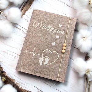 May include: A tan felt Mutterpass cover with white embroidered text and design. The design includes a heartbeat, heart, and baby footprints. A thin beige cord with three wooden beads secures the cover.