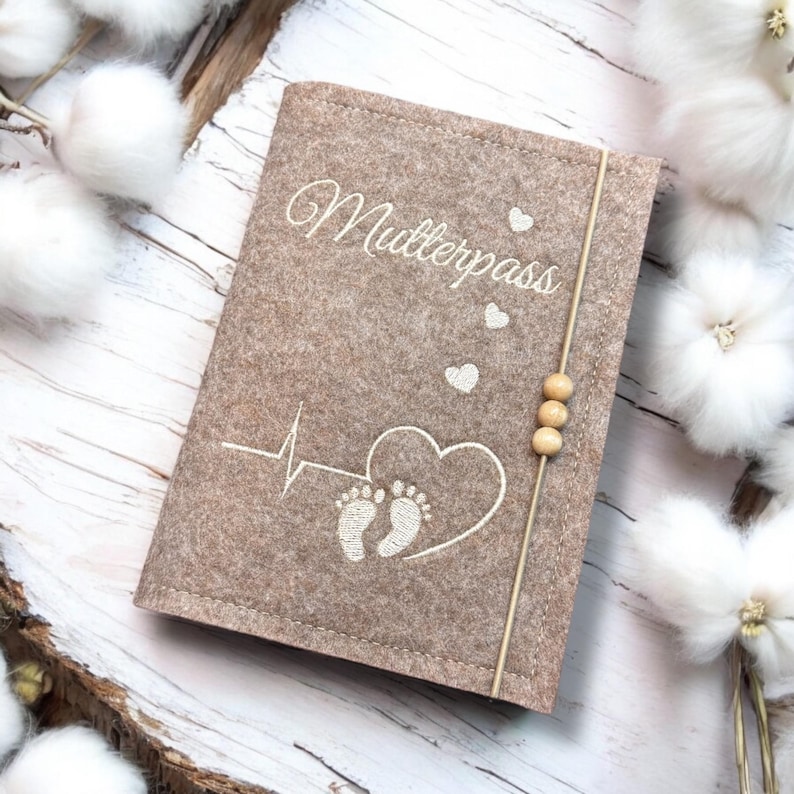 May include: A tan felt Mutterpass cover with white embroidered text and design. The design includes a heartbeat, heart, and baby footprints. A thin beige cord with three wooden beads secures the cover.