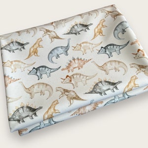 Könnte beinhalten: Gefalteter Stoff mit einem sich wiederholenden Muster aus Aquarell-Dinosauriern in Grau-, Beige- und Orangetönen auf weißem Hintergrund. Die Dinosaurier umfassen verschiedene Arten wie Triceratops und Stegosaurus.