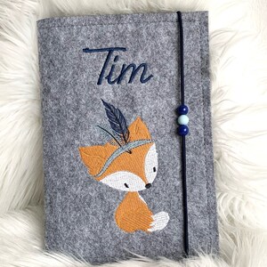 Könnte beinhalten: Grauer Filz-Buchumschlag mit dem Namen "Tim" in blauer Schrift gestickt. Mit einem orangefarbenen Fuchs mit Federkopfschmuck. Ein marineblaues Gummiband mit blauen Perlen sichert den Umschlag.