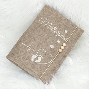 May include: A taupe felt cover with the word "Mutterpass" embroidered in white script. The cover features a heartbeat line, a heart with baby footprints, and small hearts. A wooden stick with three wooden beads is attached.