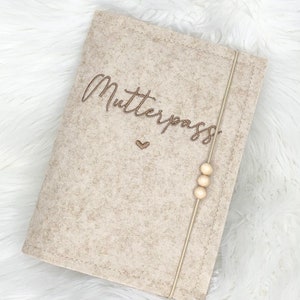 May include: A beige felt cover with the word "Mutterpass" embroidered in brown script. A small heart is embroidered below the word. A beige elastic band with three wooden beads secures the cover.