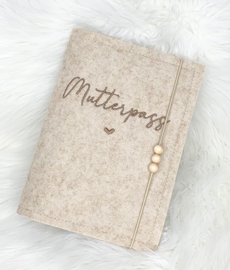 May include: A beige felt cover with the word "Mutterpass" embroidered in brown script. A small heart is embroidered below the word. A beige elastic band with three wooden beads secures the cover.
