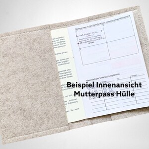 May include: A light gray felt Mutterpass cover, open to display the interior. The cover holds a document with printed text and handwritten notes. The text "Beispiel Innenansicht Mutterpass H&uuml;lle" is visible on the cover.