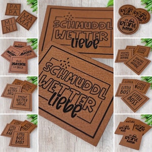 Könnte beinhalten: Eine Auswahl an braunen Lederpatches mit schwarzen Schriftzügen. Die Patches zeigen Sätze wie "SCHMUDDL WETTER liebe", "Little Boss" und "It's Beanie Season", sowie weitere Designs.